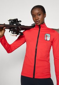 Piros sport hosszú ujjú dzseki magas gallérral és fekete cipzárral. A mellkason grafikával ellátott folt található. Síelés közben viselik.