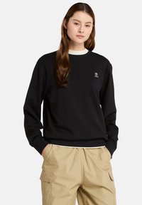Felpa nera con maniche lunghe e polsini a coste, caratterizzata da un piccolo logo. Indossata con pantaloni cargo beige.