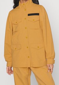 Gul jacka med hög krage, knappstängning, två fickor med klaff på framsidan och textil med struktur. Har svarta detaljer på bröstfickan.