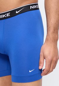 Κοντινό πλάνο της μέσης και του χεριού ενός ατόμου που φοράει μπλε εσώρουχα boxer της Nike με μαύρη μέση και λευκά λογότυπα Nike.