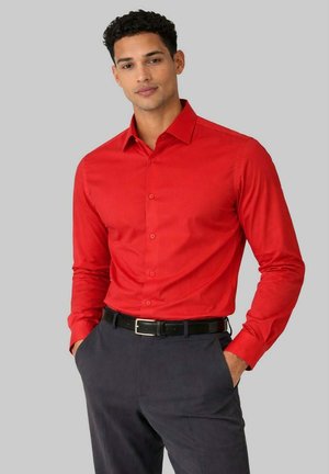 Homme aux cheveux bouclés et foncés portant une chemise rouge à manches longues ajustée avec des boutons et un pantalon gris foncé avec une ceinture noire.