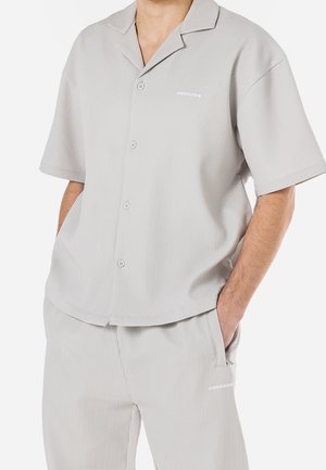 Uomo con camicia a maniche corte a coste grigio chiaro abbottonata e pantaloni abbinati, con le mani nelle tasche, su sfondo bianco.