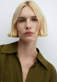 Blonde bob haircut, straight texture. Olive green shirt with a collar. Neutral background. Subtle facial freckles and natural makeup.