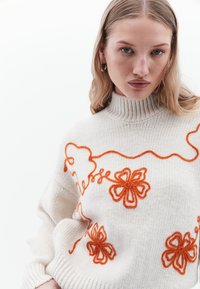 Maglione a maglia crema con colletto alto e ricami floreali arancioni. Caratterizzato da una texture a coste e una vestibilità ampia, evidenziando un design distintivo.