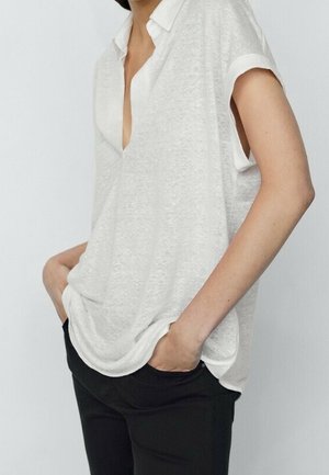 Vrouw draagt een losse witte korte mouwen blouse met kraag, lichtelijk in zwarte broek gestopt, handen in de zakken, tegen een effen lichte achtergrond.