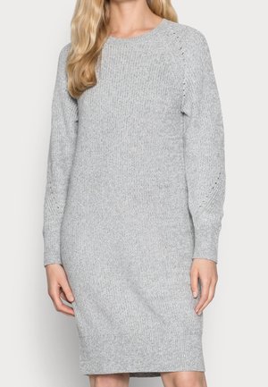 Kobieta ubrana w sięgającą kolan, jasnoszarą dzianinową sukienkę z długimi rękawami i subtelnymi ażurowymi detalami na rękawach.