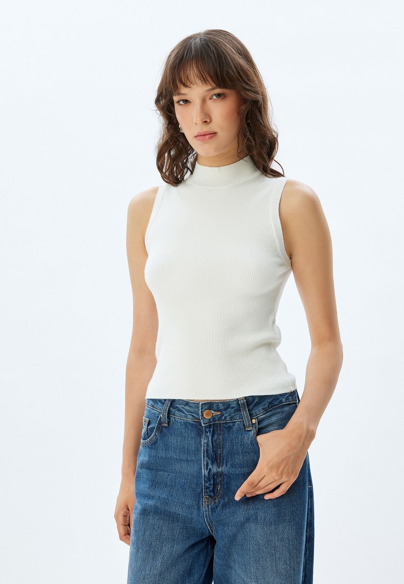 Top blanco sin mangas, de canalé y con cuello alto, combinado con jeans de tiro alto en un tono azul desgastado. Textura suave y diseño ajustado.