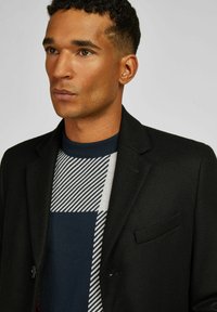 Mann mit kurzem, lockigem Haar, der einen schwarzen Mantel über einem gemusterten navy- und weißen Pullover trägt, schaut leicht nach links vor einem einfarbigen Hintergrund.