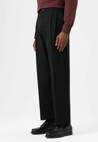 Pantalon noir plissé en tissu lisse, doté d'une coupe décontractée et d'une taille haute, associé à des mocassins en cuir noir.