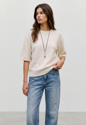 Woman wearing a cream short-sleeve sweater, blue high-waist jeans, and a long black necklace with a silver pendant, standing against a plain wall.