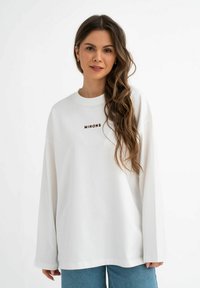 Witte sweatshirt met lange mouwen en een ronde hals, voorzien van een bruine "MIRONS" logo op de borst, gecombineerd met blauwe denim broek.