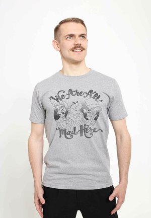 Camiseta gris con un diseño gráfico negro que presenta personajes de dibujos animados y el texto "Todos estamos locos aquí". Mangas cortas y estilo de cuello redondo.