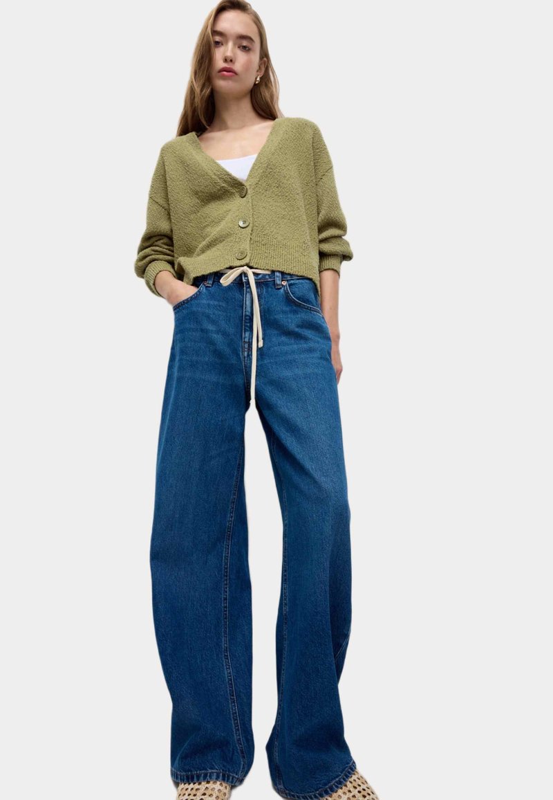 Olivově zelený pletený cardigan s výstřihem do V a knoflíky, kombinovaný s vysokými modrými džínovými kalhotami širokého střihu se stahovací šňůrkou.