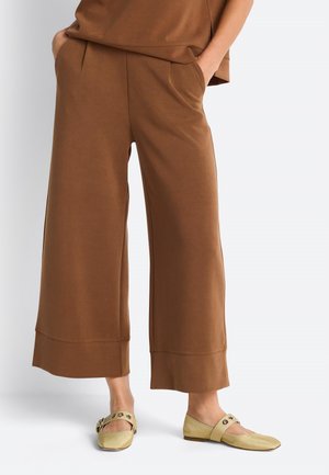 Personne portant un pantalon marron large et court avec des poches, ainsi que des chaussures plates beiges avec des brides décoratives sur un fond clair.