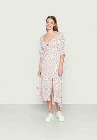 Vestido floral de comprimento médio em rosa claro, com mangas bufantes, detalhe de laço na gola e fenda lateral. Combinado com ténis brancos e uma bolsa rosa.