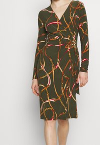 Långärmad omslagsklänning i olivgrön med ett mönster av kedja och band. Tillverkad av mjukt tyg, med en djup V-ringning och figurnära silhuett.
