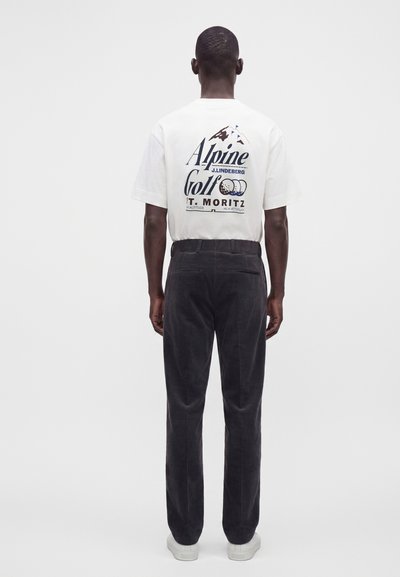 Άνδρας που φοράει λευκό T-shirt Alpine Golf και μαύρο παντελόνι, στέκεται με την πλάτη στραμμένη σε μονόχρωμο φόντο.