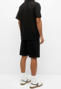 Polo shirt noir à manches courtes avec un motif tricoté texturé, associé à un short noir. Baskets blanches avec des accents noirs et beiges.