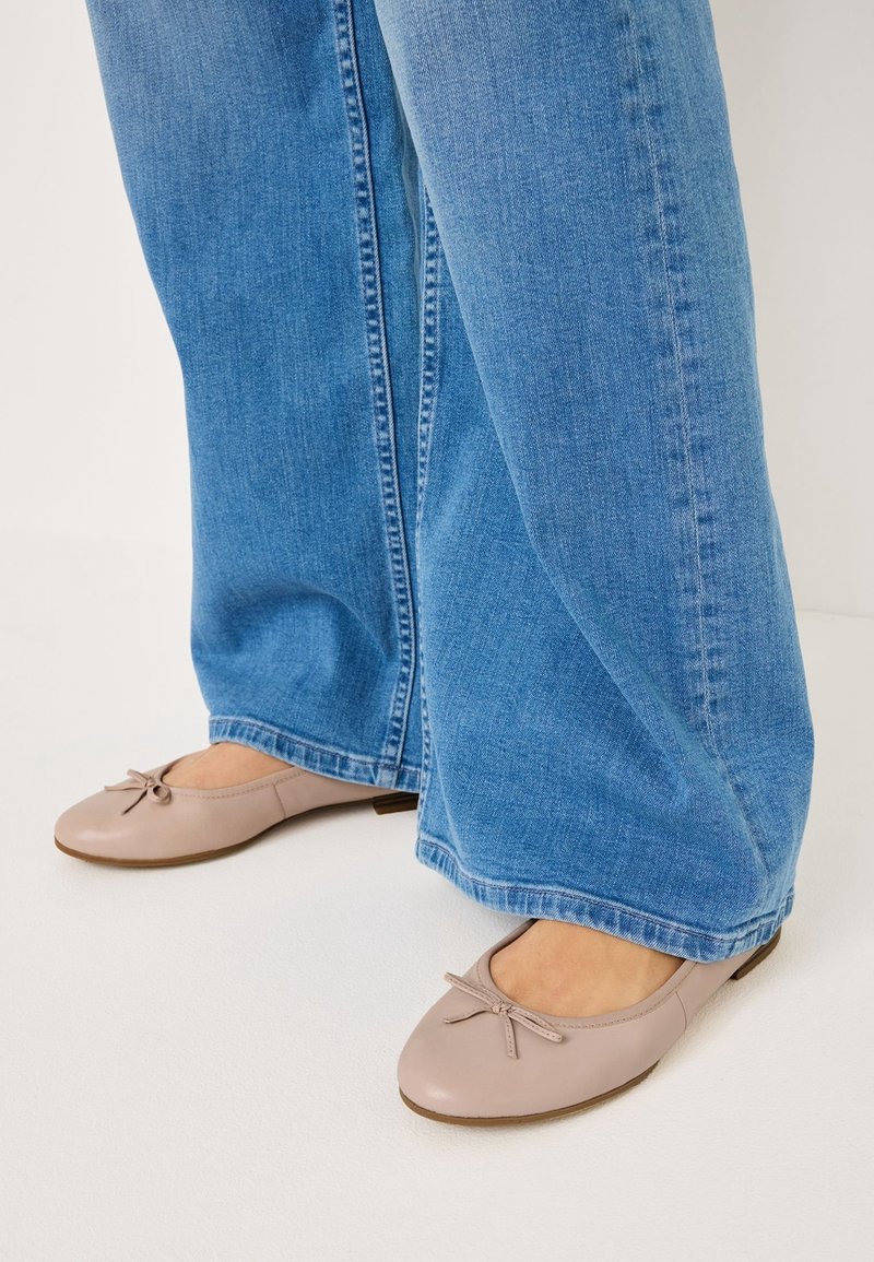 Ballerines beige clair avec petits nœuds portées avec un jean denim bleu à jambes larges sur un sol blanc.