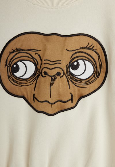 Sweatshirt de culoare crem, cu o față brodată mare, de culoare maro, cu ochi proeminenți, centrată pe piept, având o textură netedă.