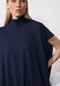 Navy blue oversized turtleneck top with short sleeves, made of smooth, stretchy material. Features a relaxed fit and draped design.
