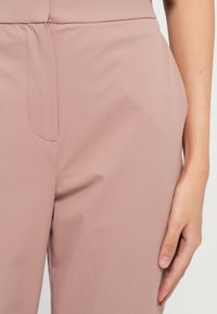 Mano destra appoggiata accanto a pantaloni a vita alta color malva con chiusura frontale nascosta e superficie in tessuto morbido.