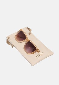Gennemsigtige amber solbriller med gradient mørke linser, der hviler på en beige pose med mærkenavnet "LIU-JO" i guld.