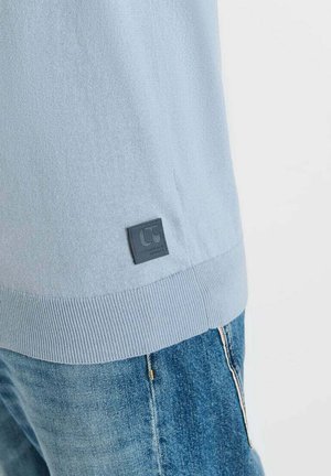Ourlet de pull en maille bleu clair avec une petite étiquette logo carrée noire, porté avec un jean en denim bleu sur un fond blanc.