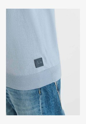 Ourlet de pull en maille bleu clair avec une petite étiquette logo carrée noire, porté avec un jean en denim bleu sur un fond blanc.