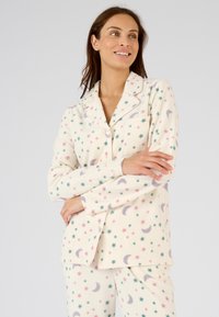 Zachte, crème kleurige pyjama set met sterren en halve maan patronen. Bevat een knoopjasje met een borstzak en een broek met elastische taille.