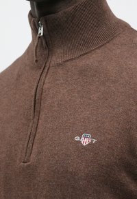 Brūns džemperis ar rāvējslēdzēju kakla daļā, izgatavots no adīta auduma, ar ribotu apkakli un nelielu GANT logotipu baltā un sarkanā krāsā uz kreisā krūšu vietā.