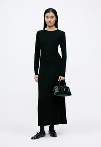 Vestido preto de manga comprida com cintura ajustada, combinado com uma pequena mala preta e sapatos pretos. Tecido liso com um design elegante.