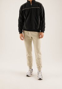 Mand iført sort zip-up sweatshirt med hvid piping, beige joggingbukser og hvide løbesko står på lys baggrund.