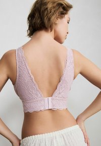 Reggiseno con schienale in pizzo di rosa pallido. Presenta un design con scollatura a V profonda bordato da un motivo ondulato e una chiusura a gancio. La texture appare delicata e intricata.