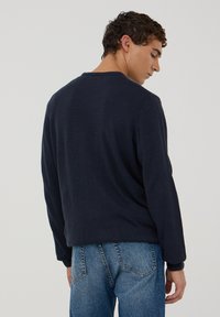 Maglione a maglia blu navy con scollo rotondo e maniche lunghe. La parte posteriore presenta una texture liscia e una vestibilità casual, abbinato a jeans azzurri.