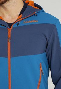 Blauwe en marineblauwe hoodiejack met oranje rits en accenten, met een zachte textuur, dubbele ritszakken en een logo op de schouder.