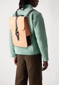 Sac à dos orange clair avec un rabat, des accents noirs et des bretelles réglables, porté sur une veste en daim vert menthe.
