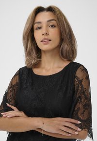 Top in pizzo nero con maniche corte a fantasia floreale. Un bracciale argentato con un'accento rosso sul polso sinistro. Capelli lisci che incorniciano il viso.