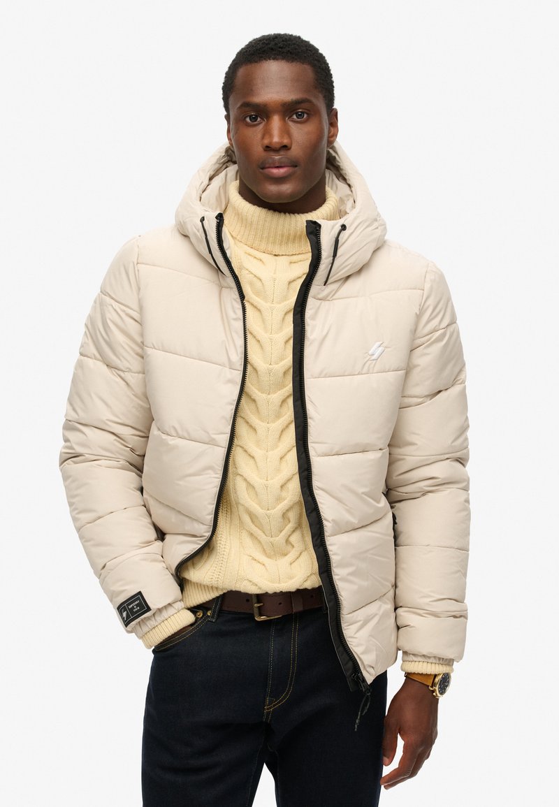 Beige Daunenjacke mit Kapuze, gesteppter Optik, schwarzem Reißverschluss und Logodetail auf der Brust, getragen über einem cremefarbenen Grobstrickpullover.