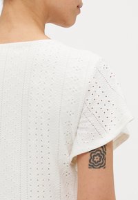 Vitt broderat linne med en texturerad design och utskurna mönster. Korta ärmar och fyrkantig halsringning, vilket visar en tatuering på armen.