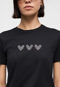 Γυναίκα που φοράει μαύρο κοντομάνικο μπλουζάκι με τρία λευκά καρδιάκια με κουκκίδες στο στήθος, καδράρισμα κάτω από τη μύτη.