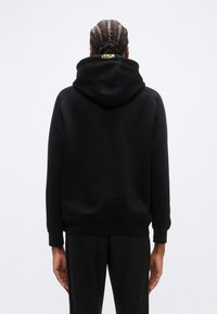 Sweat à capuche noir avec une coupe décontractée, des poignets et un ourlet côtelés. L'arrière présente un logo "VENUM" jaune au-dessus du col.