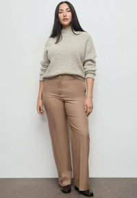 Suéter de lana beige con cuello vuelto, de corte relajado. Pantalones marrones a medida con una textura suave y pierna recta. Zapatos negros planos.