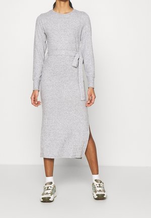 Kobieta ubrana w jasnoszary, dzianinowy, midi sweter z długim rękawem, rozcięciem z boku i pasującym paskiem do wiązania, zestawiona z białymi skarpetkami i sneakersami.