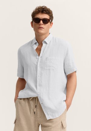 Mann trägt Schildpatt-Sonnenbrille, hellblaues kurzärmeliges Hemd mit Knöpfen und beige Cargohose mit Tunnelzug, Hände in den Taschen.