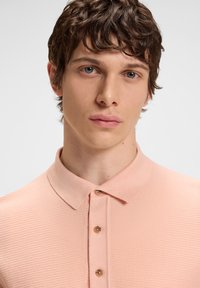 Jovem com cabelos castanho-claros e curtos, vestindo uma camisola polo texturada em pêssego claro, olhando diretamente à frente com uma expressão neutra.