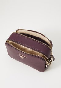 Lila Handtasche aus Kunstleder mit zwei Reißverschlussfächern, goldfarbener Hardware und einem dreieckigen Logo-Detail. Glatte Oberfläche und kompaktes Design.