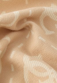 Pehmeä, teksturoitu kangas vaalean persikan sävyssä, jossa on hienovarainen valkoinen kuvio, jossa on kaartuvia muotoja ja viivoja. Materiaali vaikuttaa neulotulta ja lämpimältä.