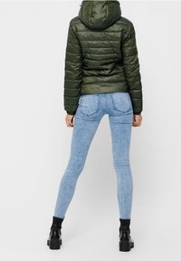 Puffed green jacket with a hood, high collar, and quilted texture paired with form-fitting light blue jeans and black ankle boots.