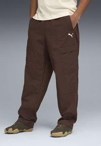 Pantalon cargo marron en tissu léger, avec poches latérales, coupe ample et petit logo blanc sur la cuisse gauche.